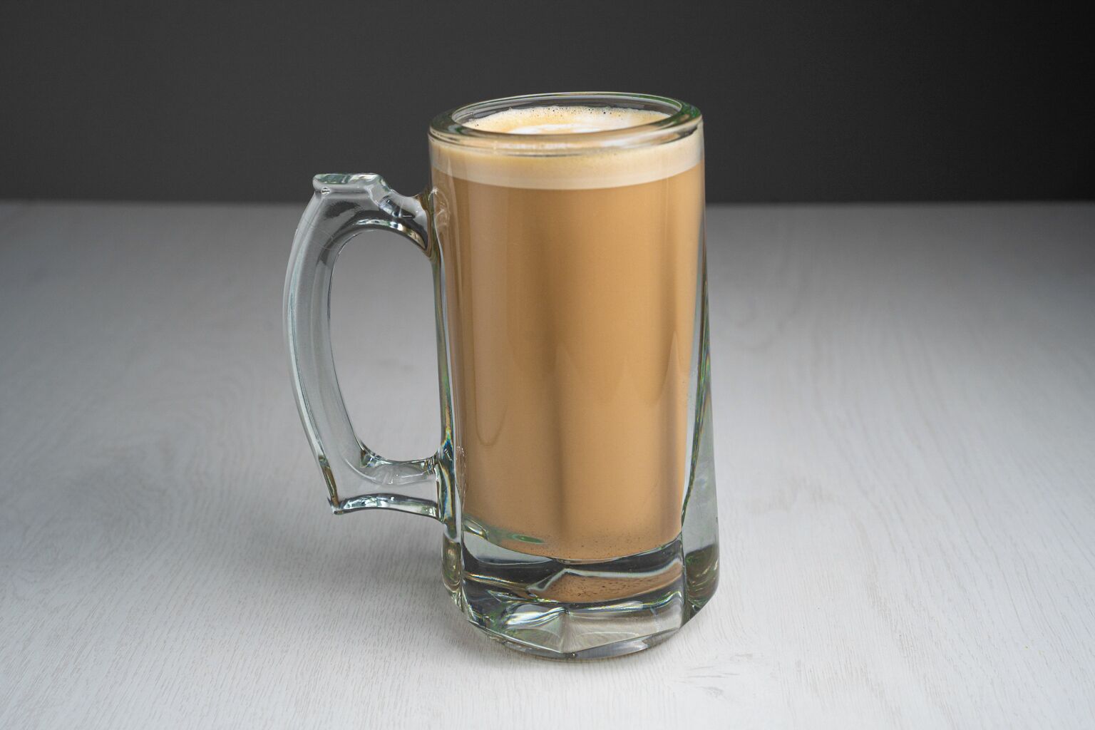 A clear glass mug with a handle, filled with a light brown beverage, placed on a light-colored surface.