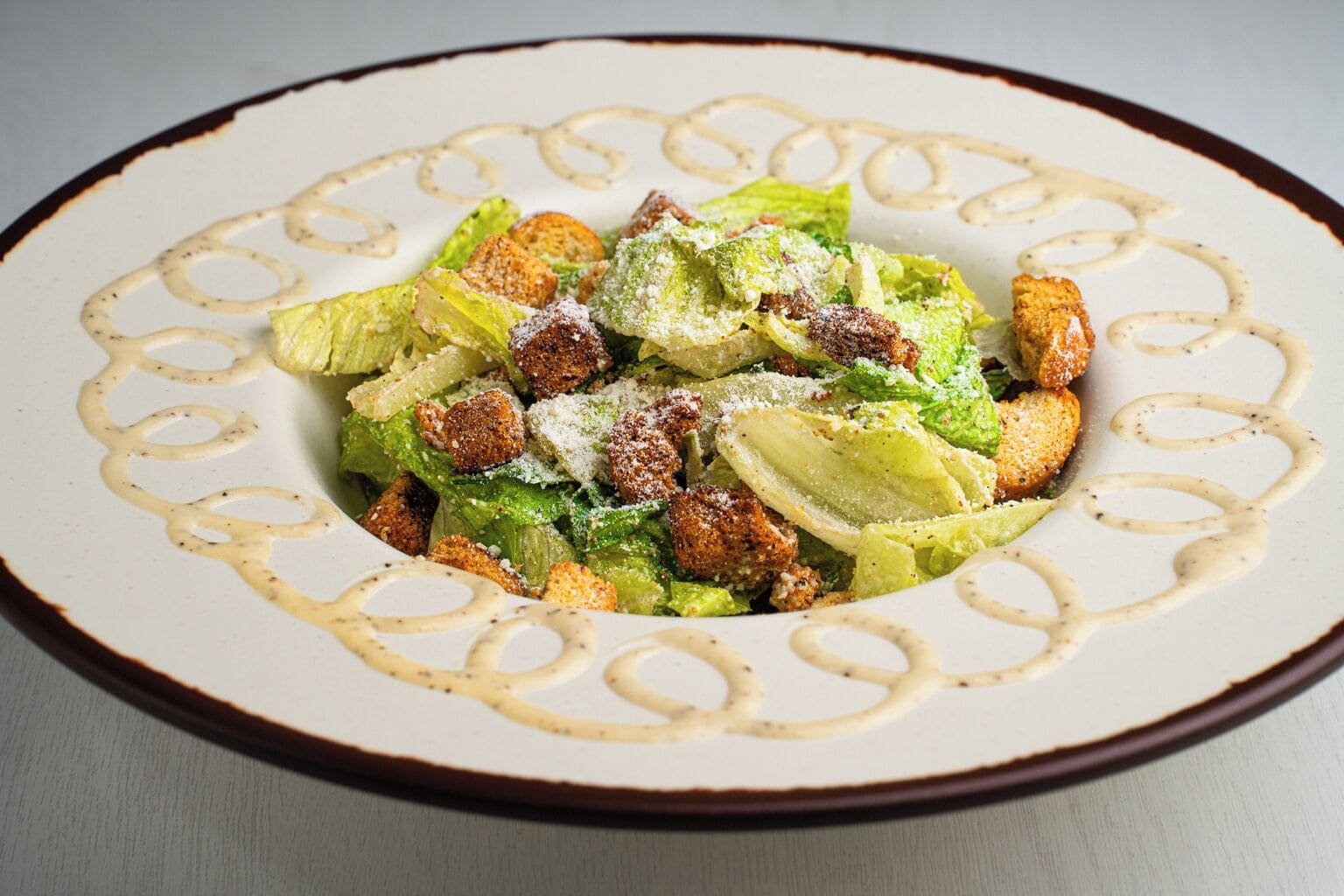A plate of Caesar salad with croutons and grated cheese, served on a decorative plate with a brown rim.