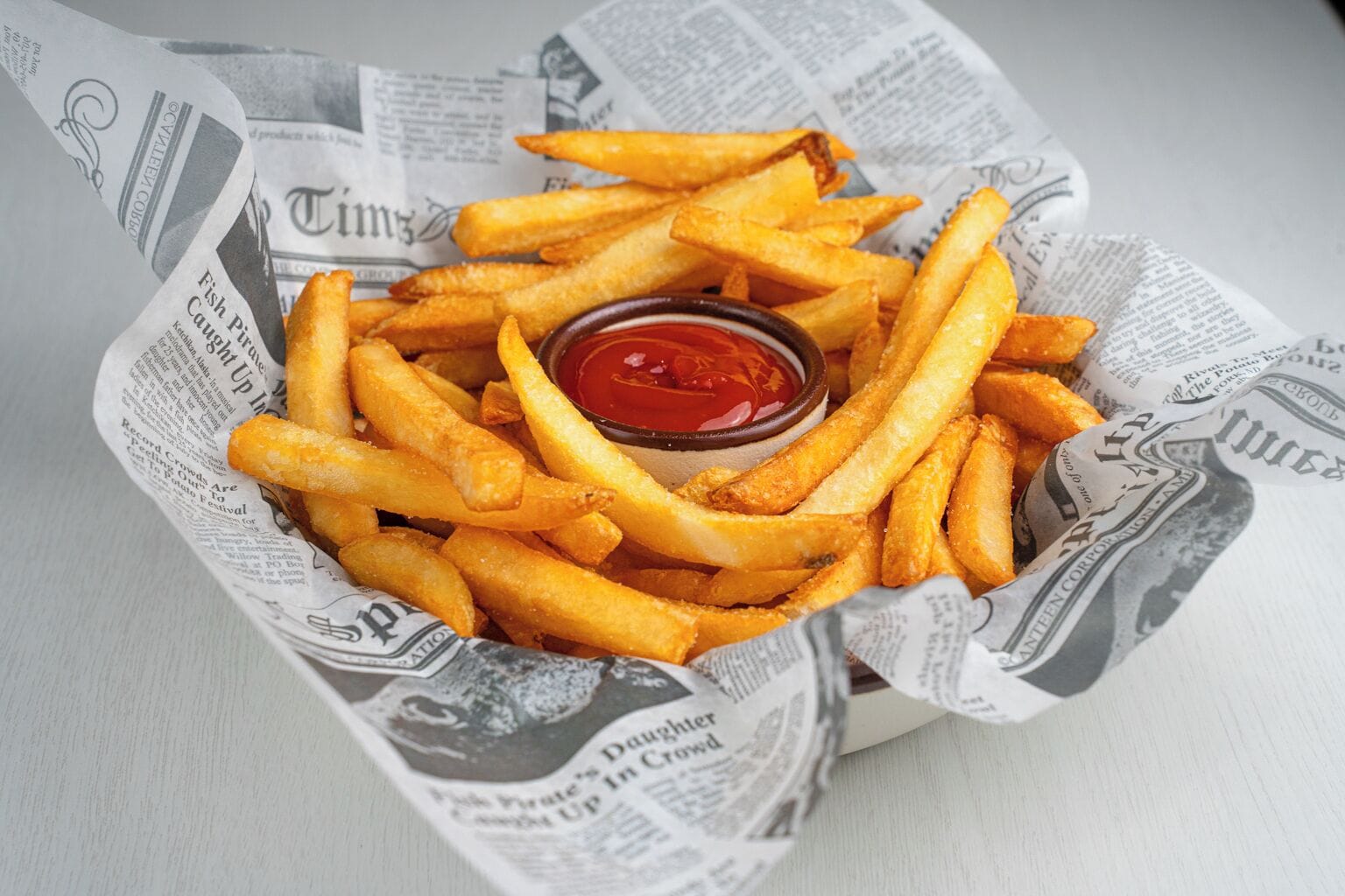 Una cesta de papas fritas doradas y un recipiente pequeño con kétchup, sobre un periódico.