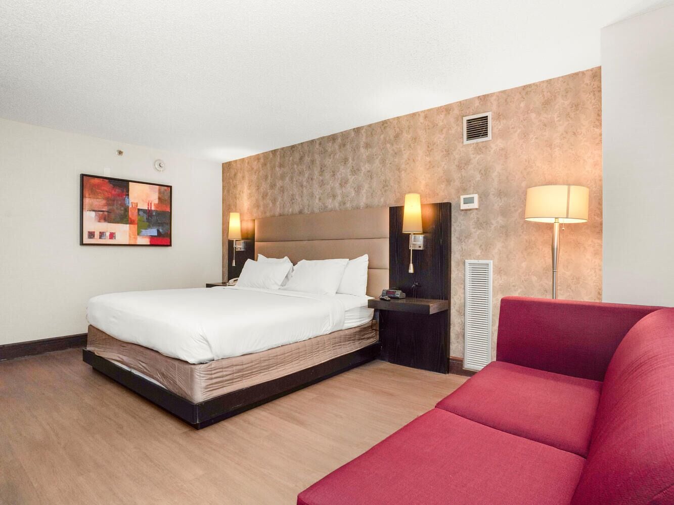 A hotel room with a bed, bedside tables, a red couch, and a wall-mounted artwork.