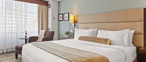Habitación de hotel con una cama grande, una silla, una mesita de noche con lámpara y una ventana con cortinas.