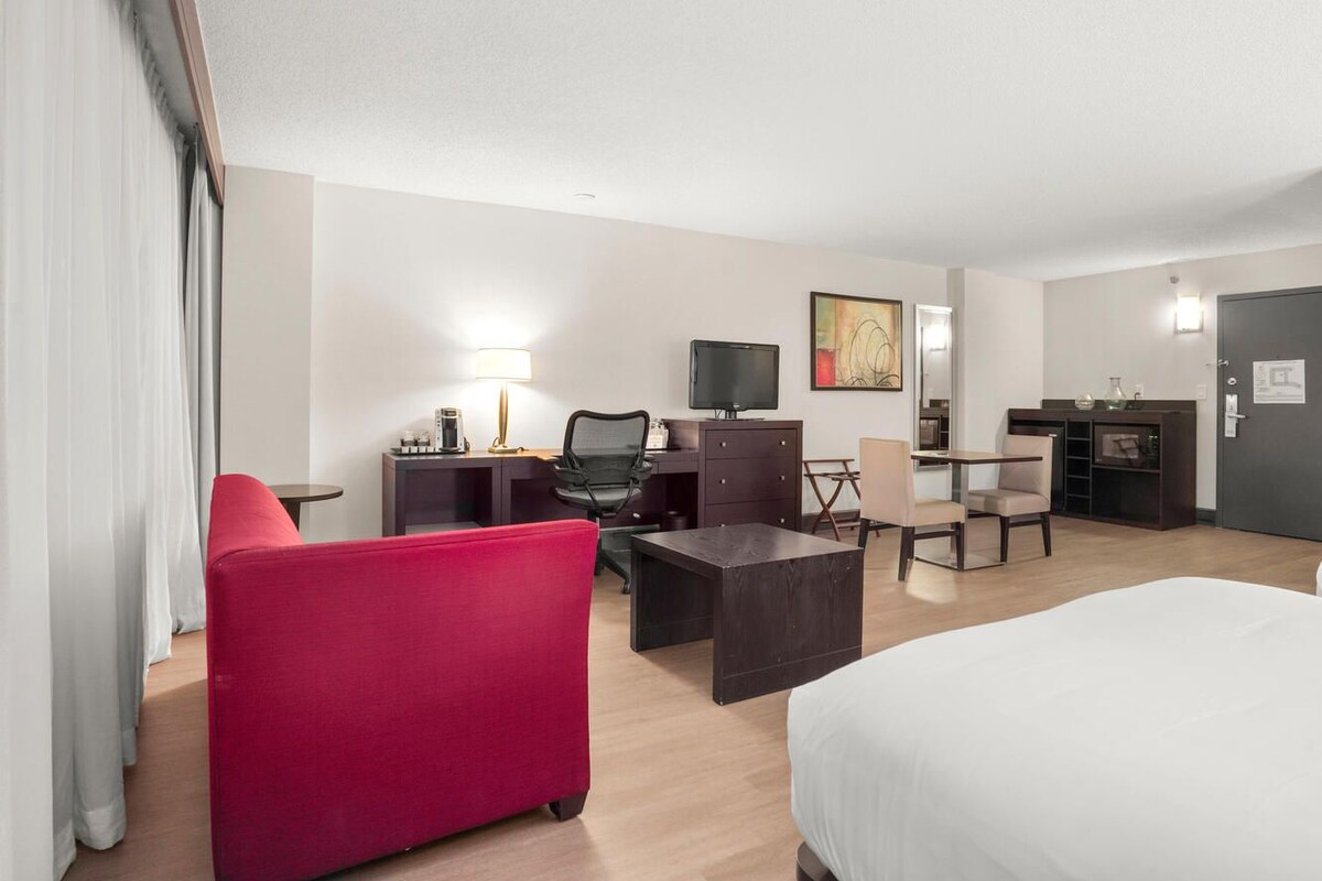 A hotel room with a bed, a desk with a computer, a chair, a red sofa, and a TV.