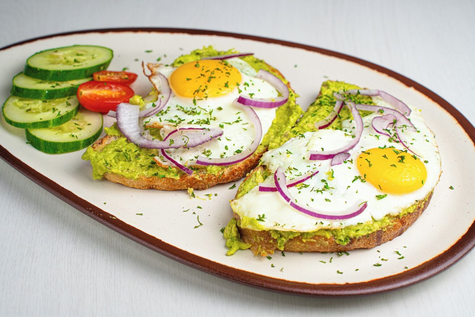 A plate with avocado toast topped with eggs and red onions, accompanied by sliced cucumbers and tomatoes.