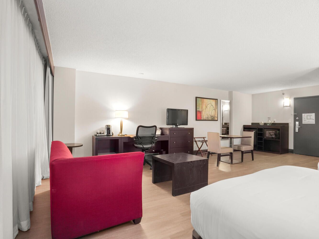 A hotel room with a bed, a desk with a computer, a chair, a red sofa, and a TV.