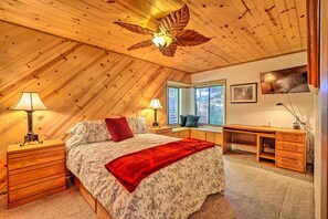 Un dormitorio con techo inclinado, una cama con ropa de cama floral, una manta roja, un escritorio con cajones y una ventana con vistas.
