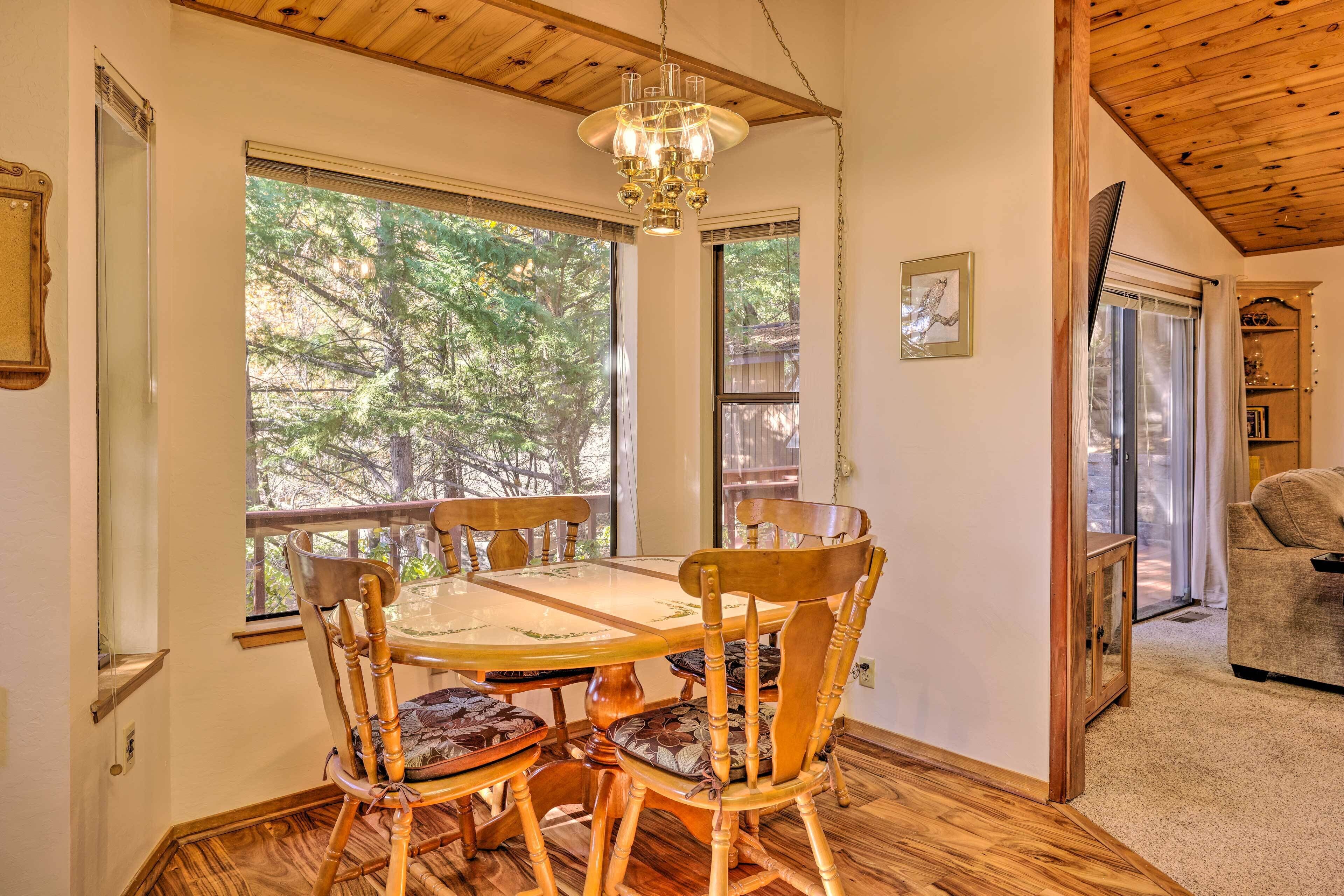 A dining area with a wooden table and chairs, large windows with a view of trees, and a chandelier.
