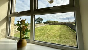 A room with a large window divided into four panes, a vase with green plants, and a hanging bird feeder. - Garden Cottage (Alnwick)