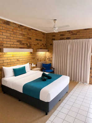 Quarto de hotel com parede em padrão tijolo, cama com roupa de cama branca e verde-água, ventilador de teto e janela com cortinas.