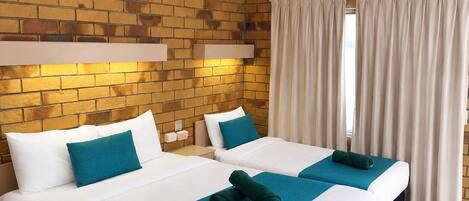 A hotel room with a brick wall, two beds with white and teal bedding, a ceiling fan, and a window with curtains.