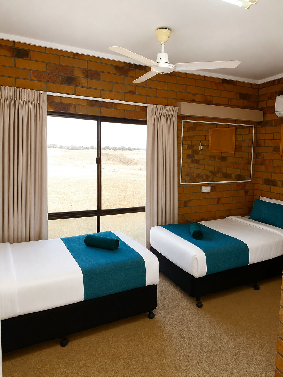 A hotel room with two beds, a ceiling fan, a large window, and a brick wall.
