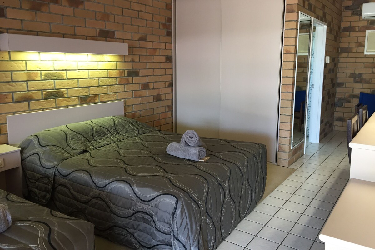 A hotel room with a brick wall, a bed with a green and gray patterned bedspread, a white bedside table, and a bathroom visible through an open door.