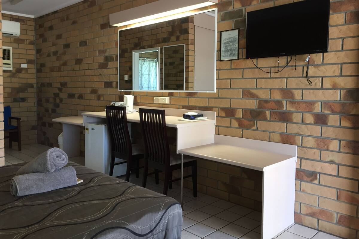 A hotel room with a bed, a desk, a chair, a mirror, a television, and a brick wall.