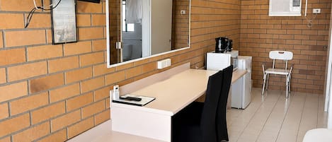 Habitación de hotel con pared de ladrillo, un televisor de pantalla plana sobre un escritorio blanco, un espejo grande, una silla blanca, una mesita pequeña blanca y una cama blanca.
