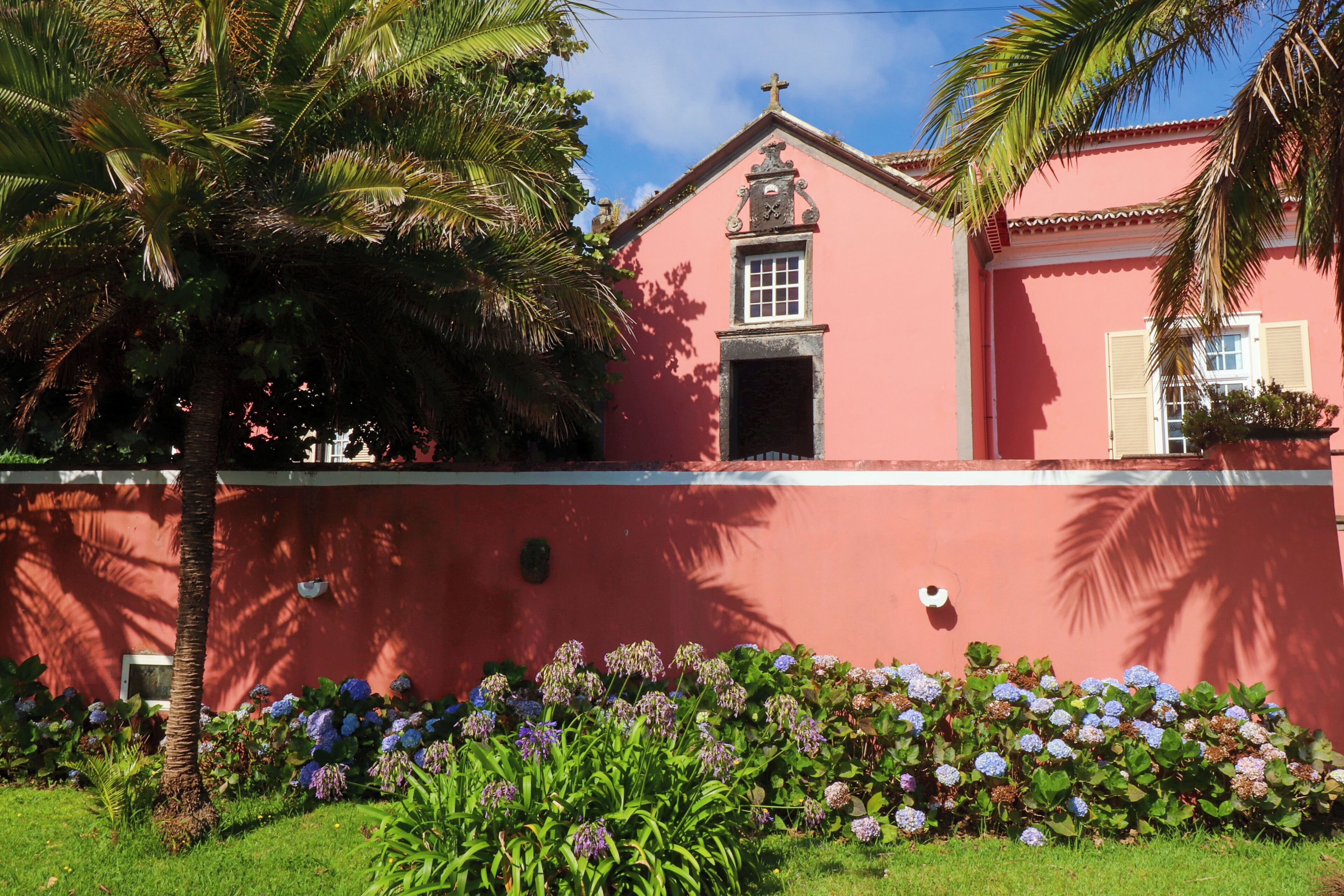 Um edifício rosa com uma cruz no topo, rodeado por um muro baixo e palmeiras.