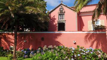 Um edifício rosa com uma cruz no topo, rodeado por um muro baixo e palmeiras.