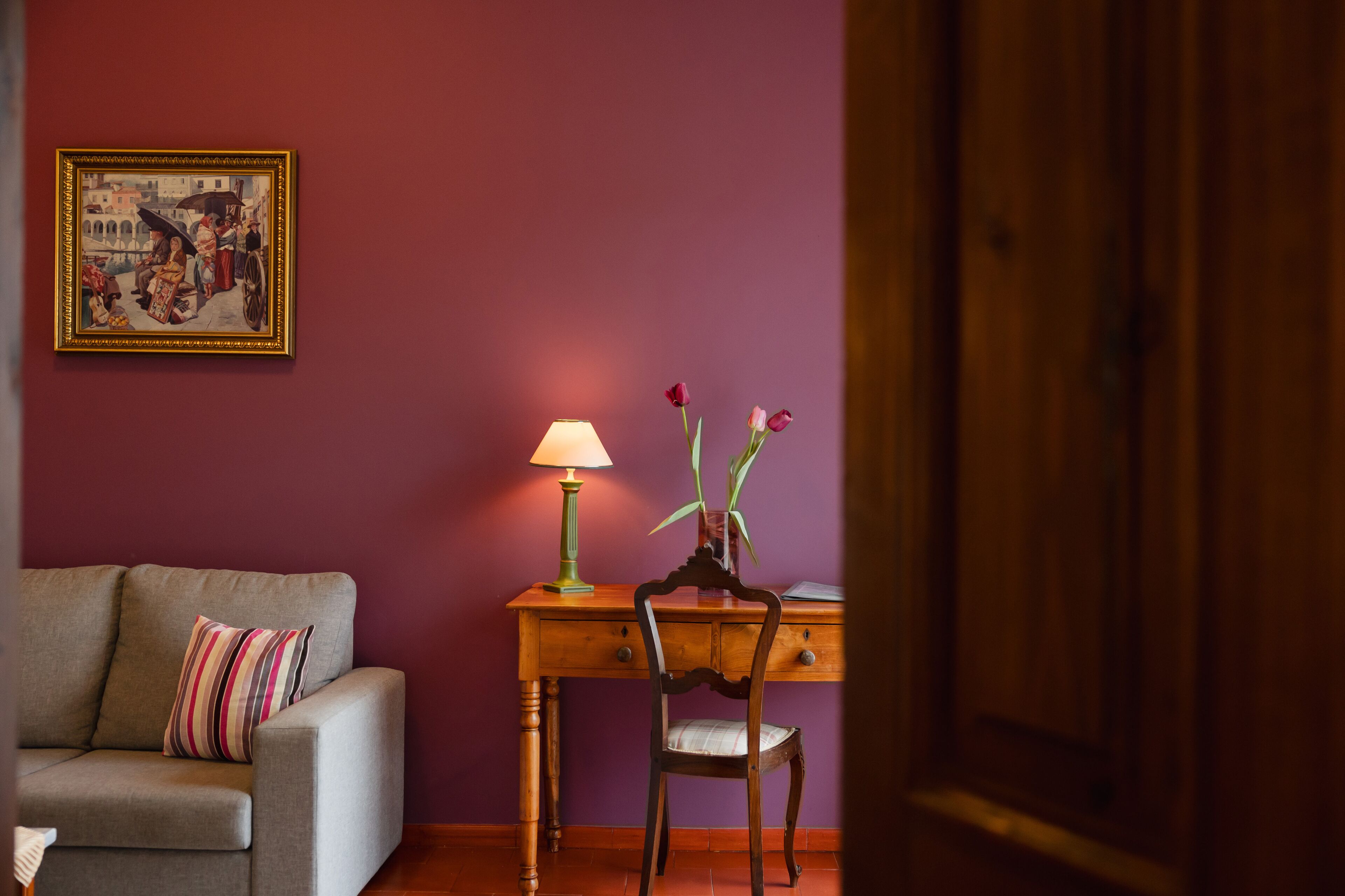A room with a sofa, a desk with a lamp, and a vase with flowers.