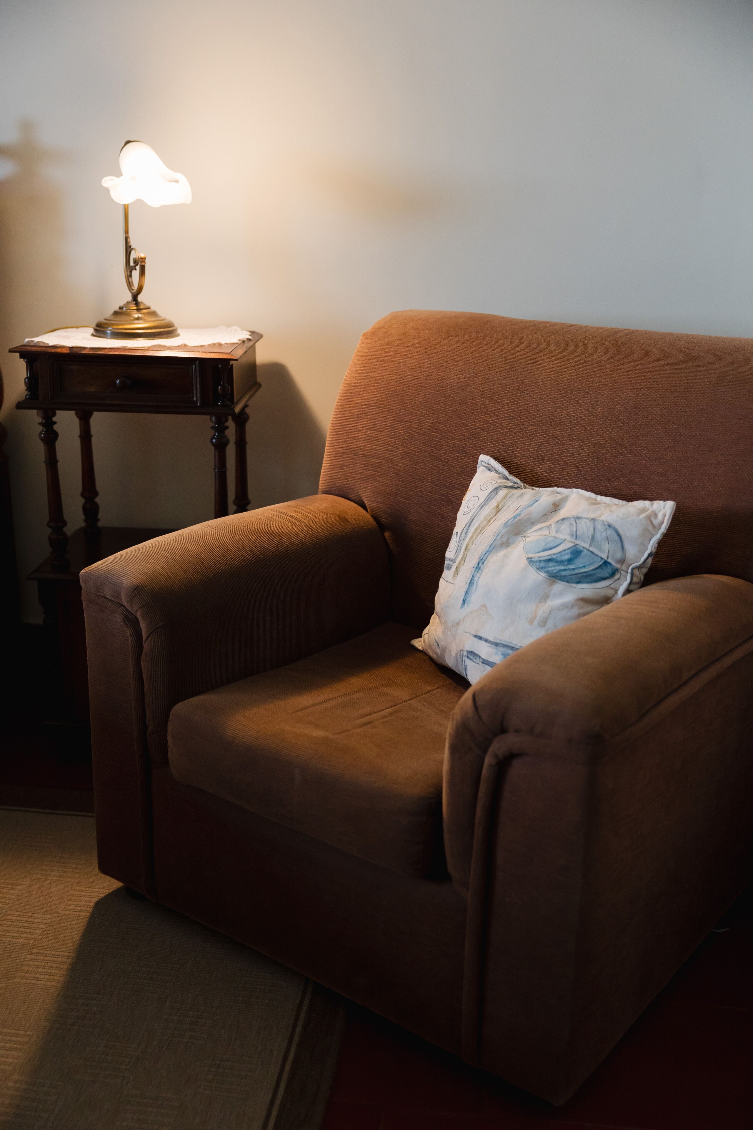 A brown armchair with a blue patterned pillow, a wooden side table with a lamp, and a rug on the floor.