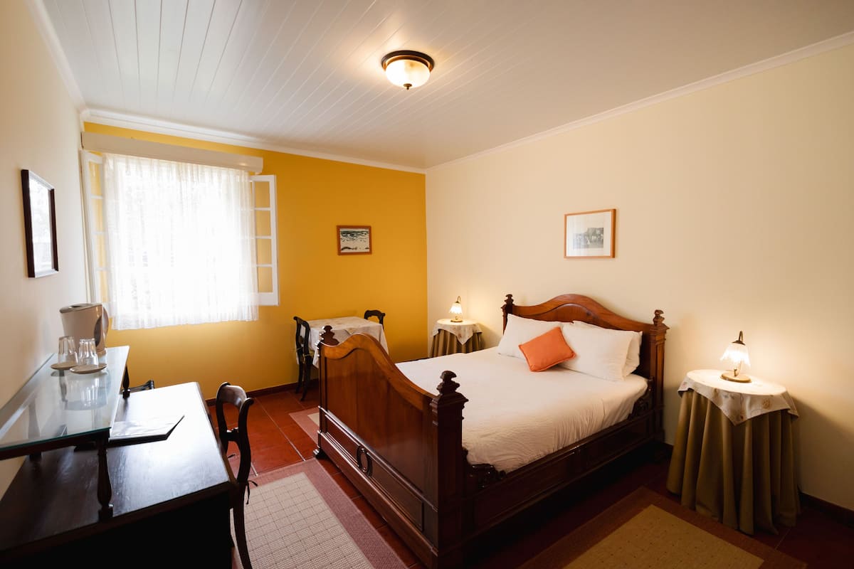 Un dormitorio con una cama de madera, un escritorio con una silla, una mesita de noche con una lámpara y una ventana con cortinas.