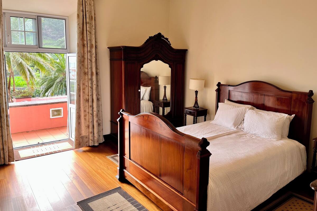 Un dormitorio con una cama de madera, una mesita de noche con una lámpara, un espejo y vistas a la vegetación exterior.