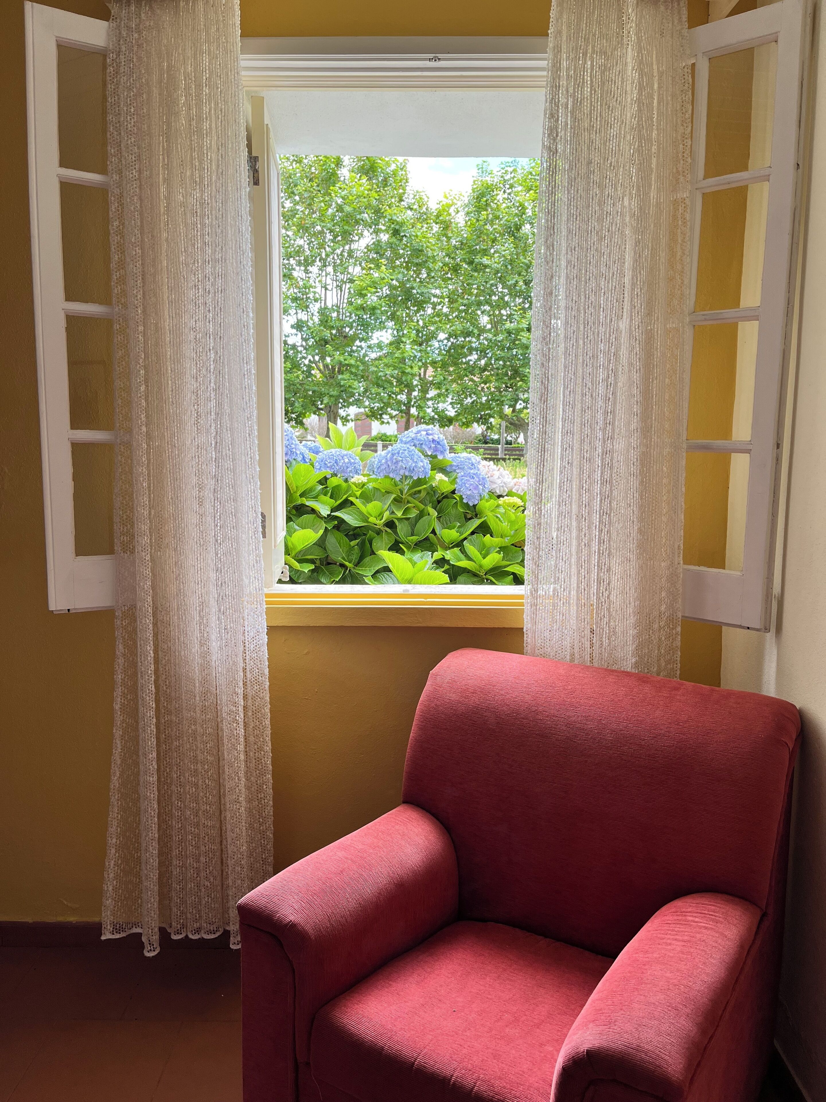 Ein Zimmer mit einem roten Sessel, einem Fenster mit weißen Vorhängen und Blick auf grüne Pflanzen und blaue Blumen draußen.