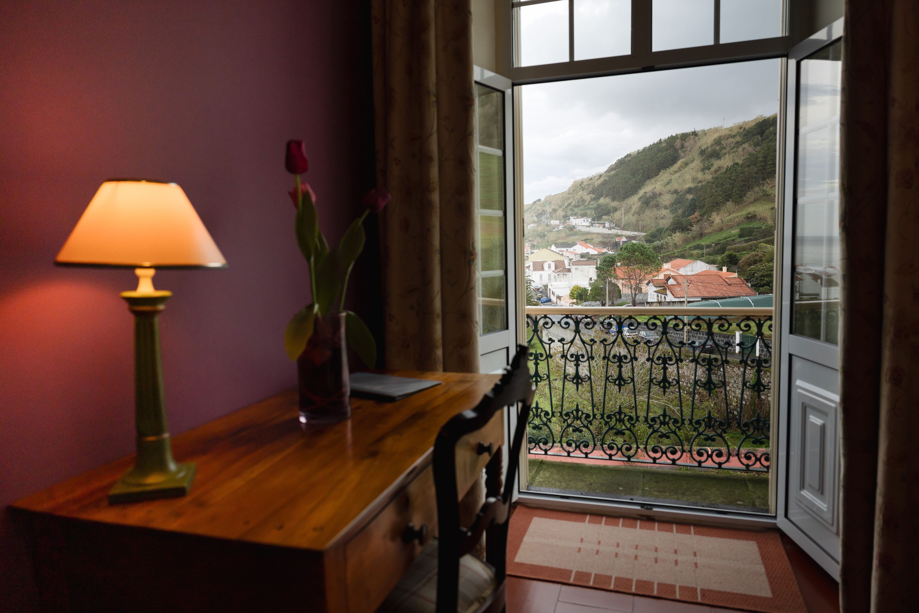A room with a wooden desk, a lamp, and a view of a town through a window with a balcony.
