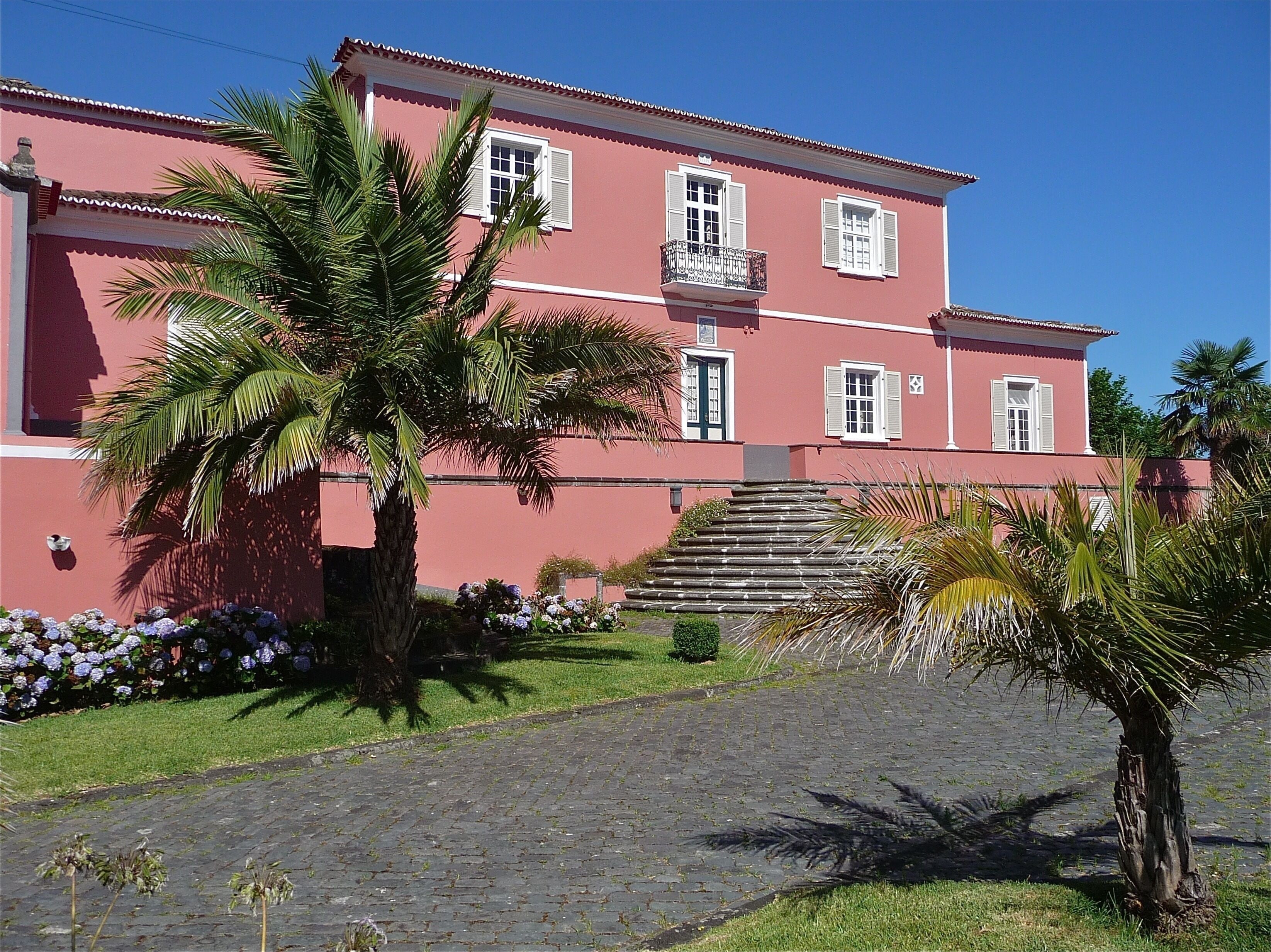 A pink building with white shutters, a balcony, and a cobblestone pathway leading to the entrance.