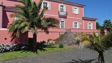 A pink building with white shutters, a balcony, and a cobblestone pathway leading to the entrance.