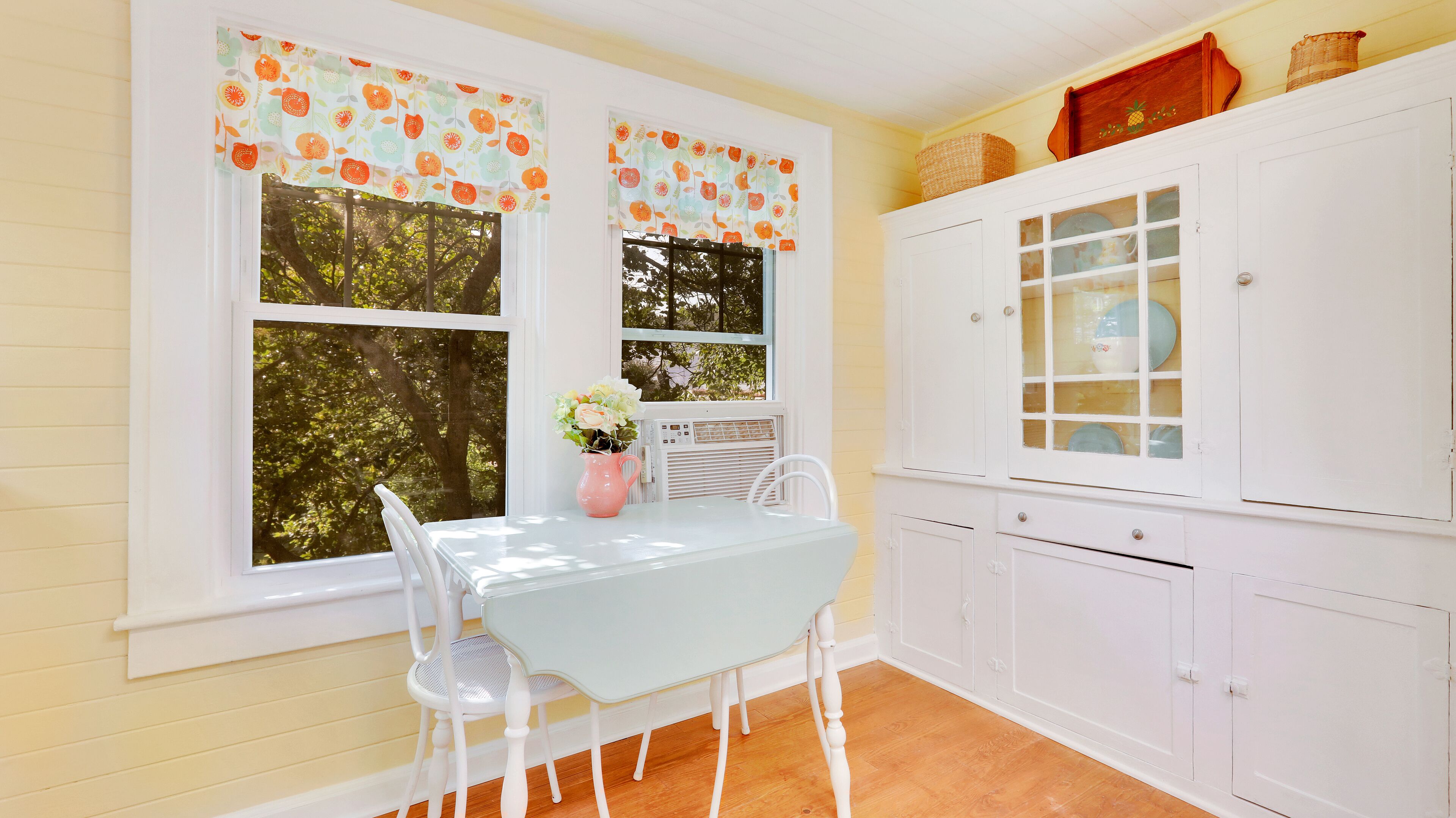 Una habitación con una mesa y sillas blancas, una ventana con un dosel floral, un armario blanco con puertas de vidrio y suelo de madera.