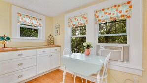 A kitchen with white cabinets, a glass table, and a window with a floral curtain. - Updated 1930s carriage house in Ardsley Park (Savannah)
