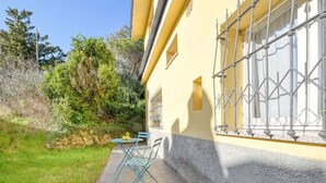 A yellow building with a stone wall and a window with metal bars. There is a small table and two chairs outside.