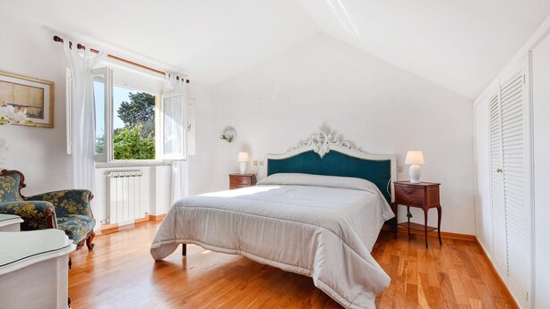 A bedroom with a large bed, a chair, a bedside table, a window with curtains, and a radiator.