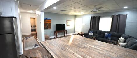 A spacious living area with a large wooden dining table, a black sofa, and a kitchen area with a refrigerator.