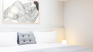 Un lit bien fait, avec une literie blanche et un oreiller gris à motifs, une œuvre d’art abstraite accrochée au mur et une lampe de chevet.