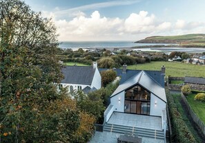 Une maison située sur la côte, dotée d’une grande baie vitrée, d’un toit en pente et offrant une vue sur la mer et les collines lointaines.