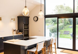 A modern kitchen with a large island, double ovens, hanging pendant lights, and a view of the garden through large sliding doors.