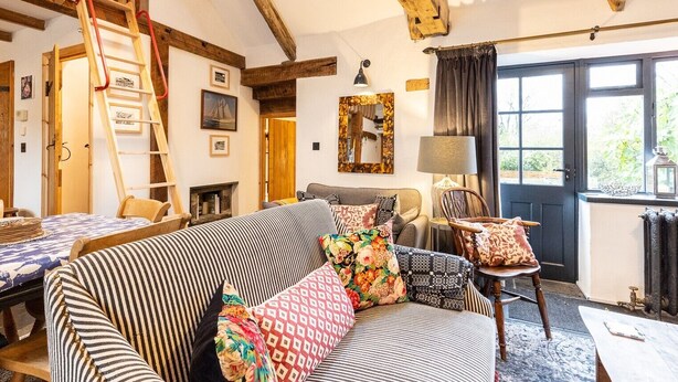 Un salon chaleureux avec une cheminée, des poutres en bois, un canapé agrémenté de coussins à motifs, une échelle et une vue par la fenêtre.