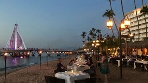 Ein Essbereich am Strand mit Tischen und Stühlen, im Hintergrund eine Brücke und Palmen.