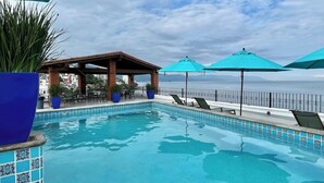 Ein Pool mit Liegestühlen und Sonnenschirmen, eine überdachte Sitzgelegenheit und Blick aufs Meer.
