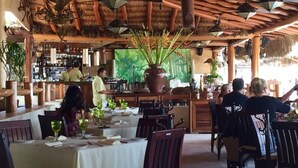 A restaurant with wooden tables and chairs, a bar area, and a large potted plant in the center.