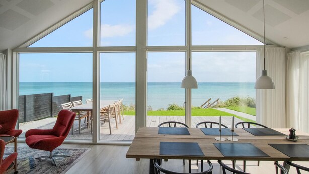 Un espacio amplio con ventanas grandes que dan al mar, una mesa de comedor de madera con sillas y un sillón rojo.