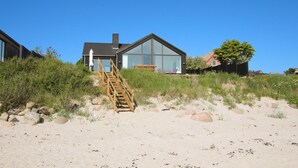 Una casa moderna frente al mar, con una escalera de madera que lleva a una puerta de cristal, rodeada de arena y vegetación.