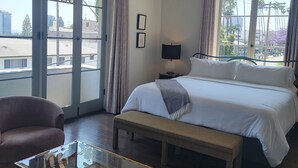Hotelkamer met een groot bed, een glazen salontafel, een bankje en uitzicht op de stad door grote ramen.