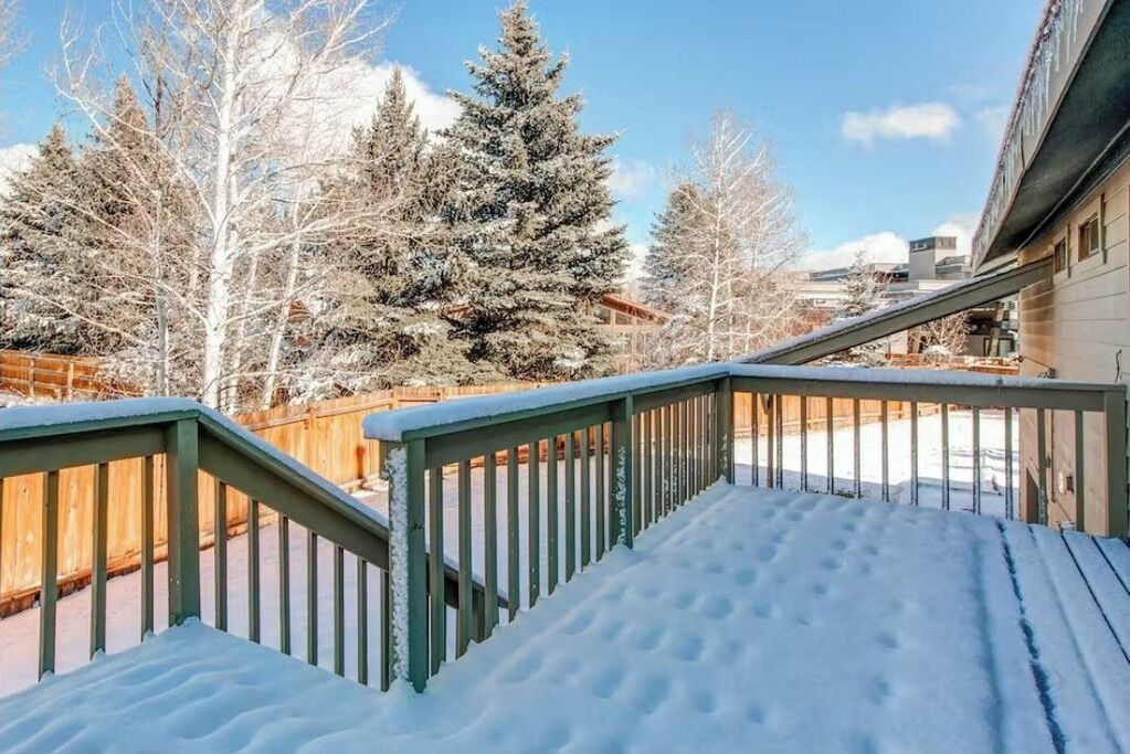 Una terraza cubierta de nieve con barandilla verde, rodeada de árboles y un edificio.