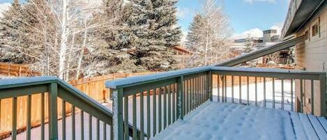 Una terraza cubierta de nieve con barandilla verde, rodeada de árboles y un edificio.