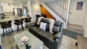 A modern living room with a black leather sofa, a coffee table, and a kitchen area with a refrigerator and bar stools. - 3BR Park Ave Townhome (Park City)
