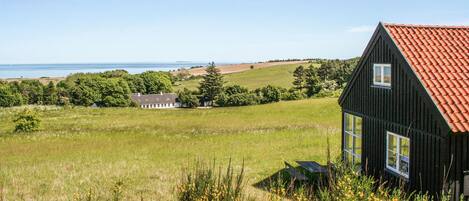 A black house with a red roof, surrounded by a grassy field and trees, with a view of the sea in the background.