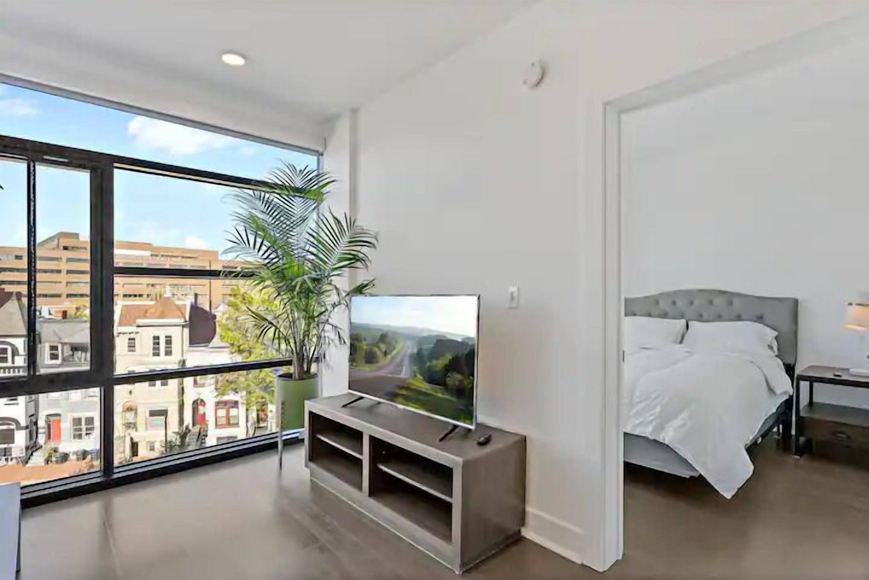 A modern bedroom with a large window, a flat-screen TV on a wooden stand, a bed with white bedding, and a potted plant.