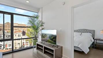 A modern bedroom with a large window, a flat-screen TV on a wooden stand, a bed with white bedding, and a potted plant.
