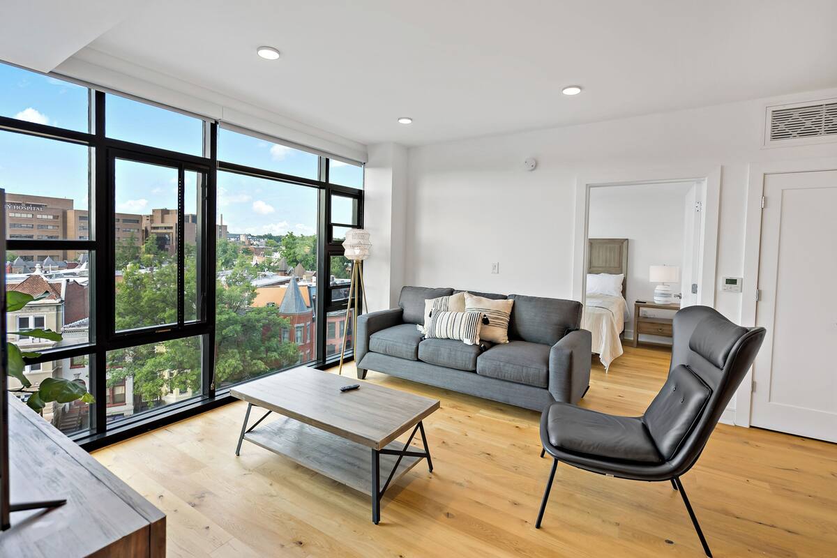 A modern living room with a large window, a grey sofa, a black leather chair, and a wooden coffee table.