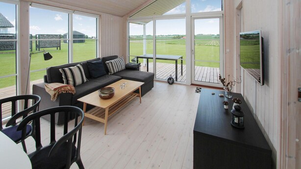 A modern living room with a black sofa, wooden coffee table, and black chairs. There is a TV mounted on the wall and a large window offering a view of a green field.